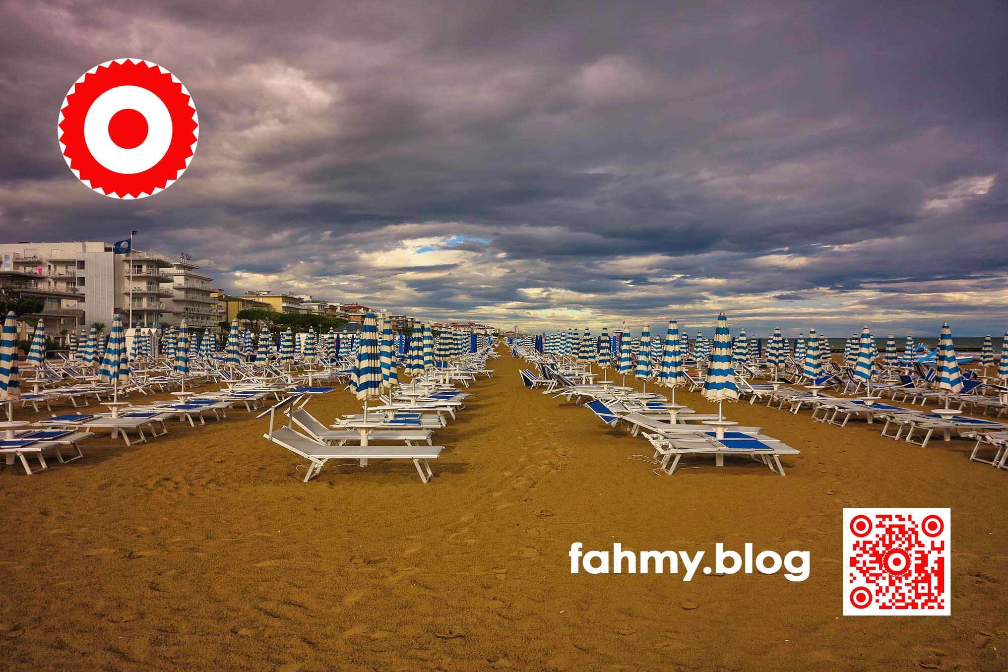 Auf dem Strandfoto sind vier ewig lange Reihen von Liegestühlen auf einem Sandstrand zu sehen. Kein Mensch ist auf dem Foto zu sehen. Über dem STrand sind dramatische Wolken zu sehen, die einen Sturm ankündigen. Rehcts oben ist das Logo von fahmy.blog - der neunundzwanzig-zackige Stern in Rot auf weißem Untergrund und angesetzt rotem Kern in der Mitte des Logos zu sehen. In der linken unteren Ecke findet sich der rot-weiße QR-Code und die Bildunterschrift "fahmy.blog".