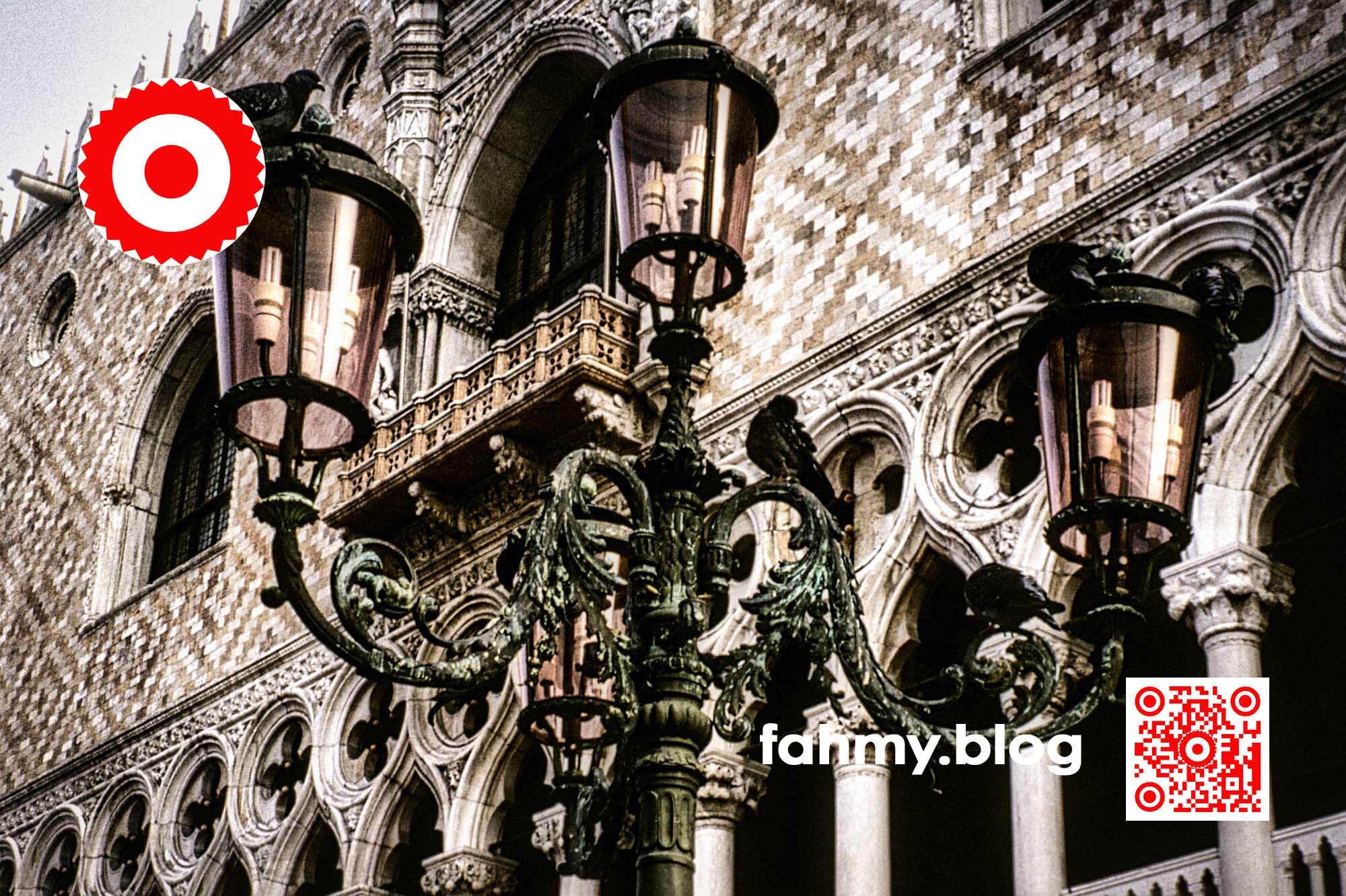 Venedig (c) 2022 fahmy.blog Venedig 2022. Der Dogenpalast beziehungsweise eigentlich nur ein Ausschnitt davon. Im Vordergrund ist eine dreiarmige Straßenlaterne in Froschperspektive - im Hintergrund die gegen San Marco gerichtete Außenwand des Dogenpalastes - zu sehen. Das rot-weiß-rote Logo von fahmy.blog ist in der linken oberen Ecke, der QR-Code und Bildunterschrift von fahmy.blog ist in der rechten Ecke zu sehen.
