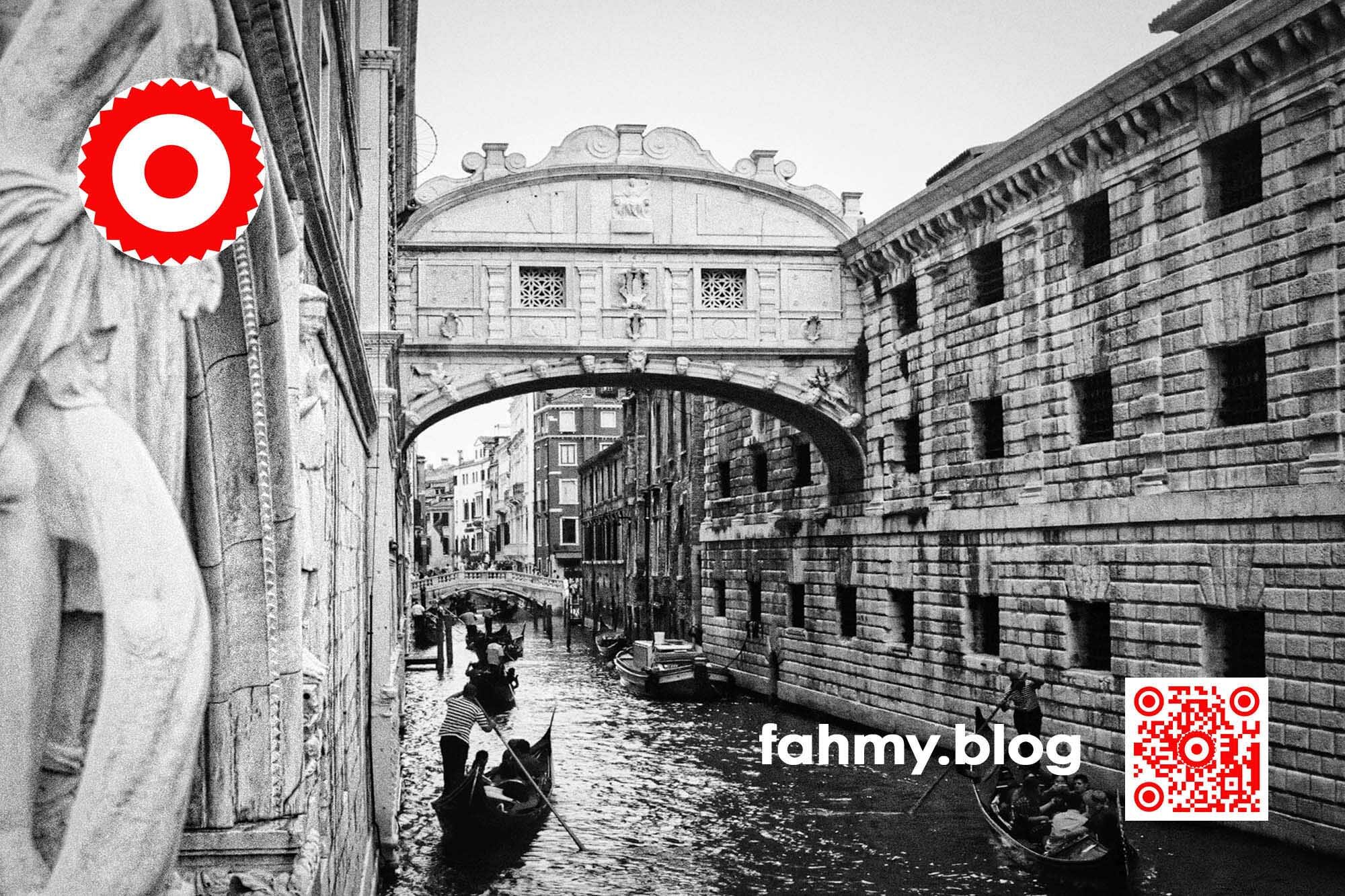 Ponte dei Sospiri (c) 2022 fahmy.blog Venedig 2022. Die Seufzerbrücke (italienisch Ponte dei Sospiri), die den Dogenpalast über einen kleinen Kanal mit dem Gefängnis verbindet. Die Seufzerbrücke inmitten des Fotos. Im Hintergrund weitere (offene) Brücken über den Kanal. Vendigkundige wissen, dass im Rücken des Fotografs die Lagune und die Vaporetto-Stationen von San Marco sind. Auf dem Kanal sind einige Gondolieri mit ihren venezianischen Gondeln zu sehen. Das Foto ist in Schwarzweiß. Gestochen scharf. Das rot-weiß-rote Logo von fahmy.blog ist in der linken oberen Ecke, der QR-Code und Bildunterschrift von fahmy.blog ist in der rechten Ecke zu sehen.