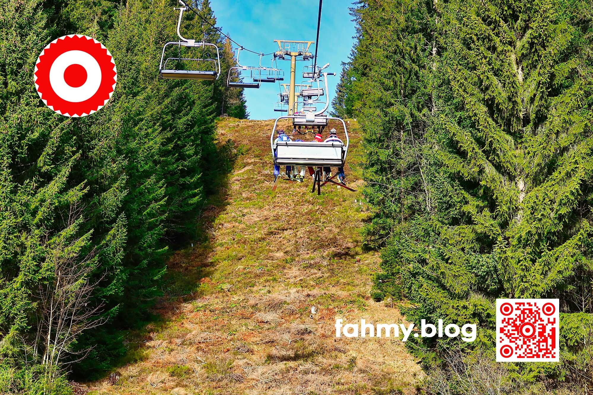 Finde den Fehler: auf dem Bild sieht man 4 Schifahrer auf einem 4er Sessellift den Berg hinauffahren. Rechts und links sind grüne Bäume und grenzen die Schneise der Lifttrasse ein. Nirgendwo ist Schnee zu sehen. Es ist ein heller, sonniger Tag. Die Schifahrer am Lift wirken in dieser Szenerie unwirklich, skurril. Das rot-weiß-rote Logo von fahmy.blog ist in der linken oberen Ecke, der QR-Code und Bildunterschrift von fahmy.blog ist in der rechten Ecke zu sehen.