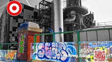 Das Bild zeigt die Müllverbrennungsanlage der Fernwärme Wien im Hintergrund - in Schwarz-Weiss - hinter einem Geländer, das im Vordergrund des Bildes mit Graffiti beschmiert ist. Der Vordergrund erscheint in grellen Farben. Auf dem Geländer sitzen zwei Tauben. Die Fassade der Müllverbrennungsanlage von dem österreichischen Künstler Friedensreich Hundertwasser gestaltet worden. Das rot-weiß-rote Logo von fahmy.blog ist in der linken oberen Ecke, der QR-Code und Bildunterschrift von fahmy.blog ist in der rechten Ecke zu sehen.