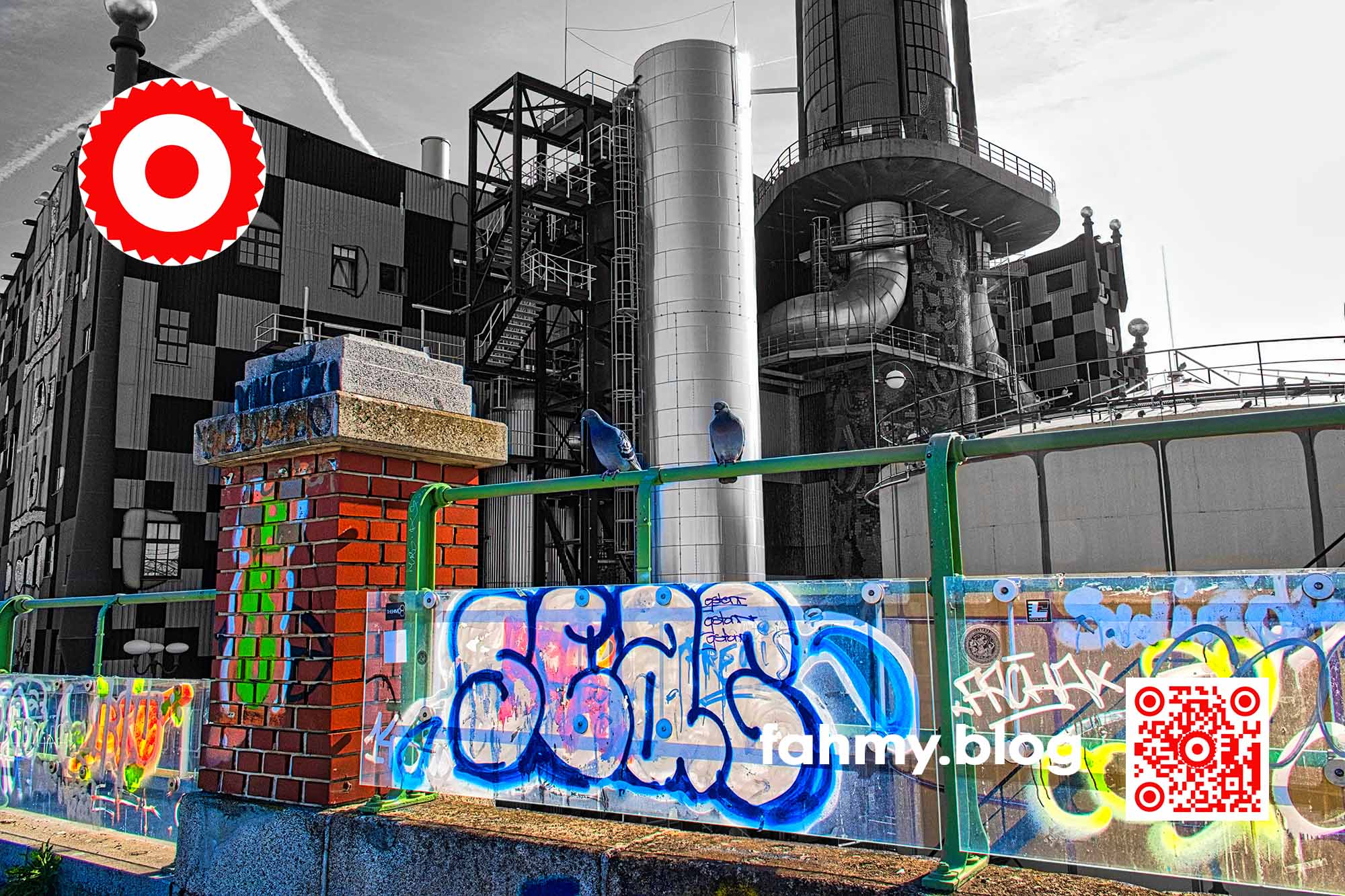 Das Bild zeigt die Müllverbrennungsanlage der Fernwärme Wien im Hintergrund - in Schwarz-Weiss - hinter einem Geländer, das im Vordergrund des Bildes mit Graffiti beschmiert ist. Der Vordergrund erscheint in grellen Farben. Auf dem Geländer sitzen zwei Tauben. Die Fassade der Müllverbrennungsanlage von dem österreichischen Künstler Friedensreich Hundertwasser gestaltet worden. Das rot-weiß-rote Logo von fahmy.blog ist in der linken oberen Ecke, der QR-Code und Bildunterschrift von fahmy.blog ist in der rechten Ecke zu sehen.