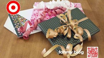 Auf dem Foto sind Geschenke und eine Glückwunschkarte zu sehen. Diese liegen auf einem Holztisch mit schöner Holzmaserung. Hell, vermutlich Kirschholz. Ein Geschenk im Vordergrund ist in dezentem schwarzem Geschenkpapier eingepackt. Auf dem schwarzen Geschenkpapier sind viele güldene kleine Kreise aufgedruckt und erinnern an ein Ornament von Klimt (oder Kettenhemd). Ein dickes ebenfalls güldenes Geschenkband ziert das Geschenk im Vordergrund. Die goldnenen Schlaufen des Geschenkbandes ziehen die Aufmerksamkeit der Betrachter/innen auf sich. Das Geschenk im Vordergrund liegt auf einem zweiten Geschenk. Dieses ist in rosarotem Geschenkpapier verpackt auf dem viele kleine rote Herzen zu sehen sind. Zwei Geschenkbänder - eines in Rosa das zweite in Weiß, werfen viele kleine Schlaufen. Das rot-weiß-rote Logo von fahmy.blog ist in der linken oberen Ecke, der QR-Code und Bildunterschrift von fahmy.blog ist in der rechten Ecke zu sehen. Unter beiden Geschenken liegt ein geöffnetes weißes Kuvert, aus dem eine Grußkarte/Geburtstagskarte, ebenfalls in Schwar mit vielen kleinen bunten Herzen bedruckt, herausblitzt.